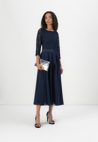 Robe A ligne marine avec un corsage en dentelle, des manches trois-quarts et une taille en satin. Le modèle porte une pochette métalisée argent et des talons noirs.