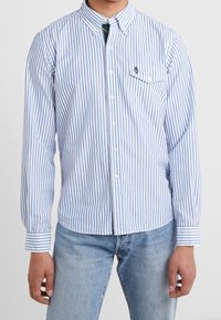 Ljust blå och vit randig skjorta med knappar, med en bröstficka, krage med knappar och lång ärm. Tillverkad av lätt tyg.