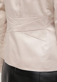Veste beige ajustée avec des détails de couture diagonale portée sur une jupe en cuir noire, vue de dos de la taille jusqu'au milieu des hanches.