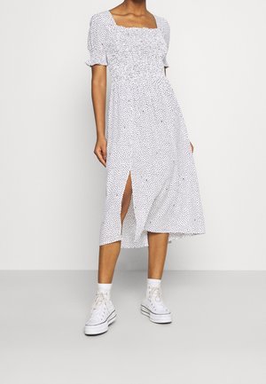 Žena v bílé midi šatech s černými puntíky, balonovými rukávy, předním rozparkem, bílých ponožkách a bílých vysokých teniskách.