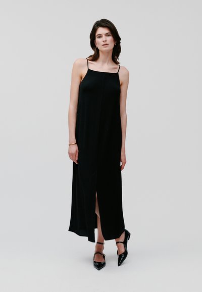 Schwarzes Maxikleid mit Spaghettiträgern, glatter Textur und einem Schlitz vorne. Getragen mit schwarzen spitzen High Heels und schlichten Schmuckakzenten.