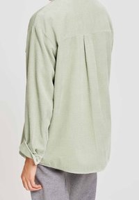 Persona che indossa una camicia di corda verde chiaro, ampia, con maniche lunghe e schiena plissettata, abbinata a pantaloni grigi, vista da dietro.