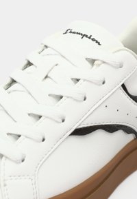 White leather sneaker with black accents, featuring rounded toe, lace-up design, textured surface, and brown rubber sole.