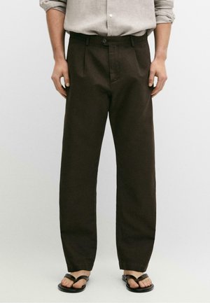 Homme portant un pantalon plissé marron foncé, une chemise gris clair retroussée et des tongs noires, debout devant un fond uni.
