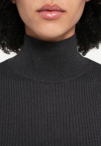 Tummanharmaa poolopaita, jonka pinta on resorikuvioitu. Lähikuva kauluksen muotoilusta, joka on istuva ja koristeltu hienovaraisilla ompeleilla.