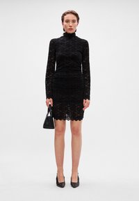 Μαύρο φόρεμα δαντέλας με μακριά μανίκια και ψηλό λαιμό, με διακοσμητική φάσα στο τελείωμα. Το μοντέλο κρατά μια μικρή μαύρη τσάντα και φοράει μαύρες μύτες.