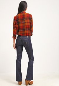 Rote und orange karierte Bluse mit hochgekrempelten Ärmeln, kombiniert mit dunkelblauen Schlagjeans. Holz-Plateau-High-Heels runden das Outfit ab.