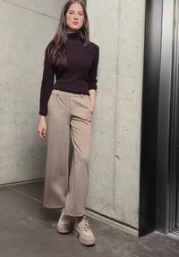 Jeune femme en pantalon beige à jambes larges, pull noir à col roulé et chaussures beiges, debout près d’un mur en béton près d’un ascenseur.