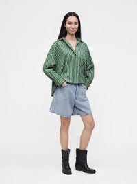 Femme aux longs cheveux noirs portant une chemise rayée verte, un short en jean bleu clair et des bottines noires, debout les mains dans les poches.