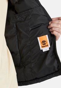 En hand håller en svart puffjacka öppen, vilket avslöjar en etikett med en trädlogotyp och varumärkesnamnet "Timberland" broderat i orange.