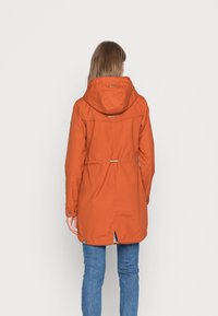 Pomarańczowa parka z kapturem, zwężona w talii, z detalami na zatrzask. Posiada wentylację z tyłu oraz miękki, teksturowany materiał. Noszona z niebieskimi dżinsami.