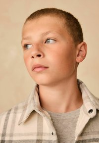 Camisa de cuadros gris y blanco con plisados y una textura suave, superpuesta sobre un suéter de cuello redondo gris claro, que cuenta con un collar abotonado.