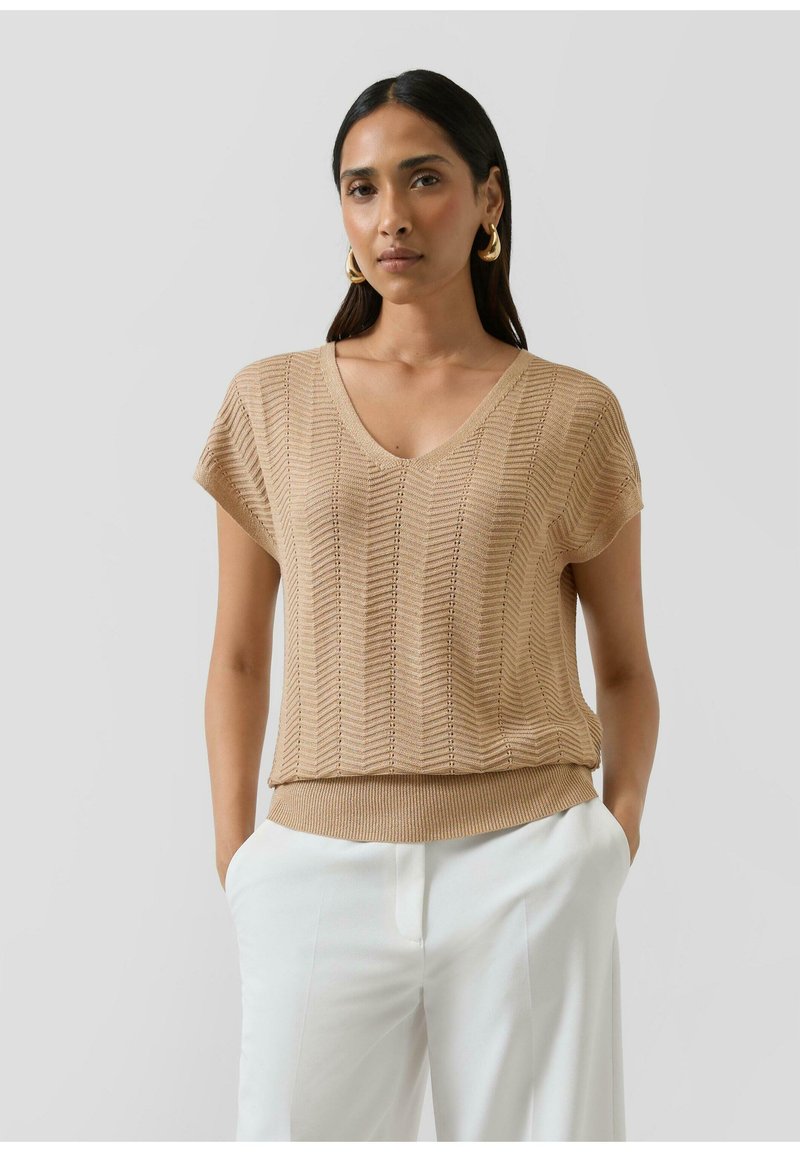 Woman wearing beige short-sleeve knit top with V-neck and white pants, standing with hands in pockets against a plain background.
