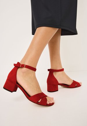 Red suede mid-heel sandals feature an ankle strap and crossed front bands. The heel is block-shaped with a textured finish.