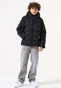 Claro abrigo de plumas negro con capucha y cierre frontal, con textura acolchada y bolsillos laterales. Combinado con jeans grises claros y zapatillas blancas.