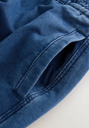 Nahaufnahme von blauen Hosen mit elastischem Bund, die eine sichtbare seitliche Tasche und detaillierte Nähte auf dem Stoff zeigen.