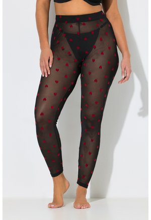 Frau trägt schwarze transparente Leggings mit roten Herzmustern, kombiniert mit schwarzer Unterwäsche und einem schwarzen, bauchfreien Top, steht barfuß an einer weißen Wand.
