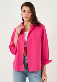 Fuchsia button-up shirt with rolled sleeves, made of lightweight fabric. Worn over a white crop top and paired with blue jeans.