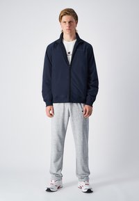 Navy lightweight jacket with zip front, ribbed cuffs, and elastic hem, paired with grey athletic pants and white sneakers.