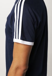 Tmavě modré bavlněné tričko s bílými pruhovanými akcenty na ramenou a bílým lemem. Design je přiléhavý a má krátké rukávy.