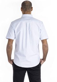 Camisa azul clara de manga corta con cuello abotonado, textura suave y pliegue trasero para un ajuste cómodo, combinada con pantalones negros.