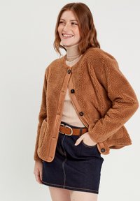 Veste en polaire marron avec des bordures en faux cuir beige et six boutons noirs, portée sur un pull col roulé beige à côtes et une jupe en jean noir.