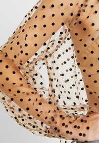 Gros plan d'un tissu transparent couleur nude avec des pois noirs couvrant les bras et le torse nus, montrant les détails de texture et de motif.