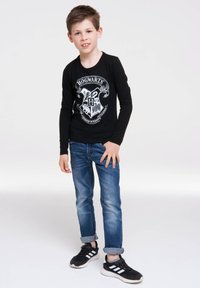 Schwarzes Langarmshirt mit einem Hogwarts-Wappen-Design, kombiniert mit blauen Jeans und schwarzen Sneakers mit weißen Streifen.