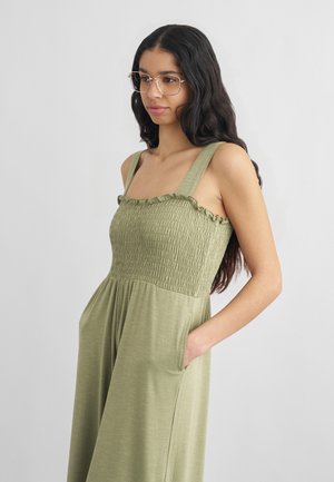 Mladá žena s dlouhými tmavými vlasy, která nosí brýle a je oblečená v bezrukávových olivově zelených šatech s nařaseným živůtkem a volánkovým okrajem, má ruku v kapse.