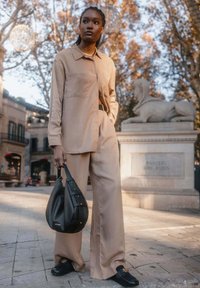 Beige Hemd mit Knöpfen, weite beige Hose, schwarze Handtasche mit Nietenakzenten und schwarze Slipper. Urbane Außenszene.
