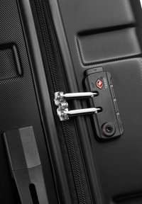 Valigia nera con zip in metallo e tirante, dotata di lucchetto approvato TSA e guscio testurizzato. Materiale liscio e resistente con finitura opaca.