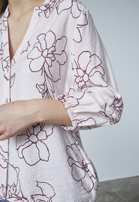 Lyserød skjorte med knapper og maroon blomsterkontur mønster, afslappet pasform og elastiske manchetter. Struktureret stof med synlige sømme.