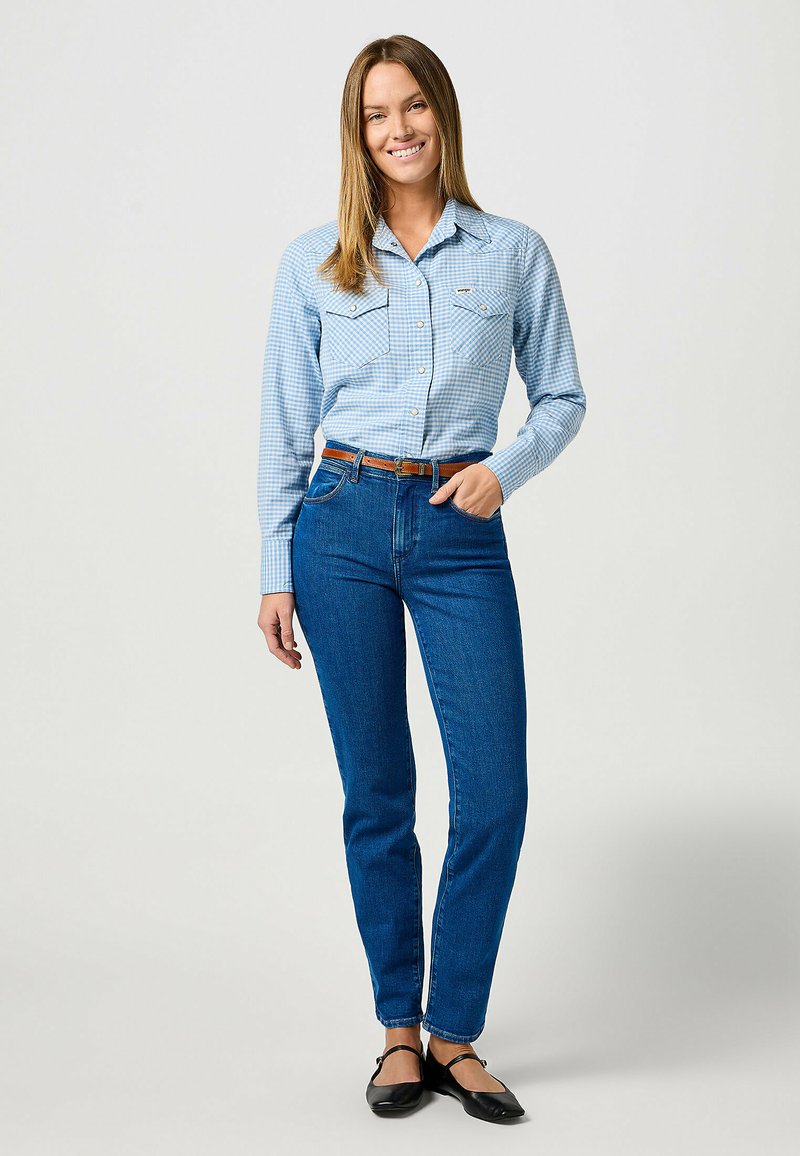 Camisa a cuadros azul claro con mangas largas y dos bolsillos en el pecho, combinada con jeans ajustados azul oscuro y zapatos negros planos, complementada con un cinturón marrón.