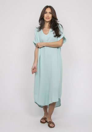 Femme aux longs cheveux bruns portant une robe midi bleu clair ample et des sandales marron, debout devant un fond clair uni.