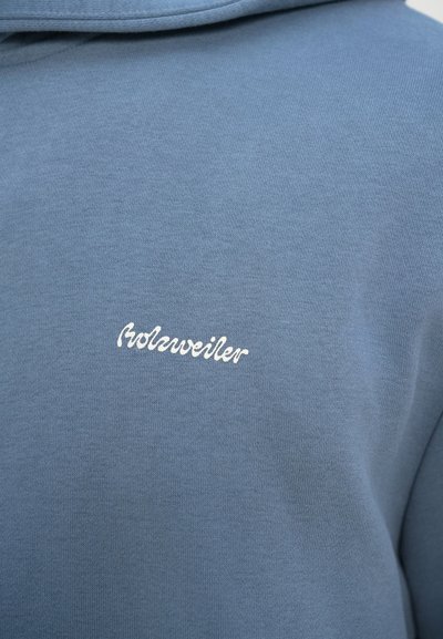 Gaiši zila kapuce ar mīkstu tekstūru un baltu izšūtu logo "rolauer" uz krūtīm, uzsverot minimālistisku dizainu.