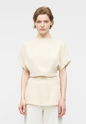 Kobieta o krótkich jasnobrązowych włosach, ubraną w kremowy top z krótkim rękawem z wiązaniem w talii oraz dopasowane jasne spodnie, stojąca na jednolitym tle.