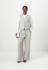 Gilet taillé gris clair avec des boutons et une petite poche, associé à un pantalon large plissé. Chemise blanche en dessous, tenant un petit sac à main.