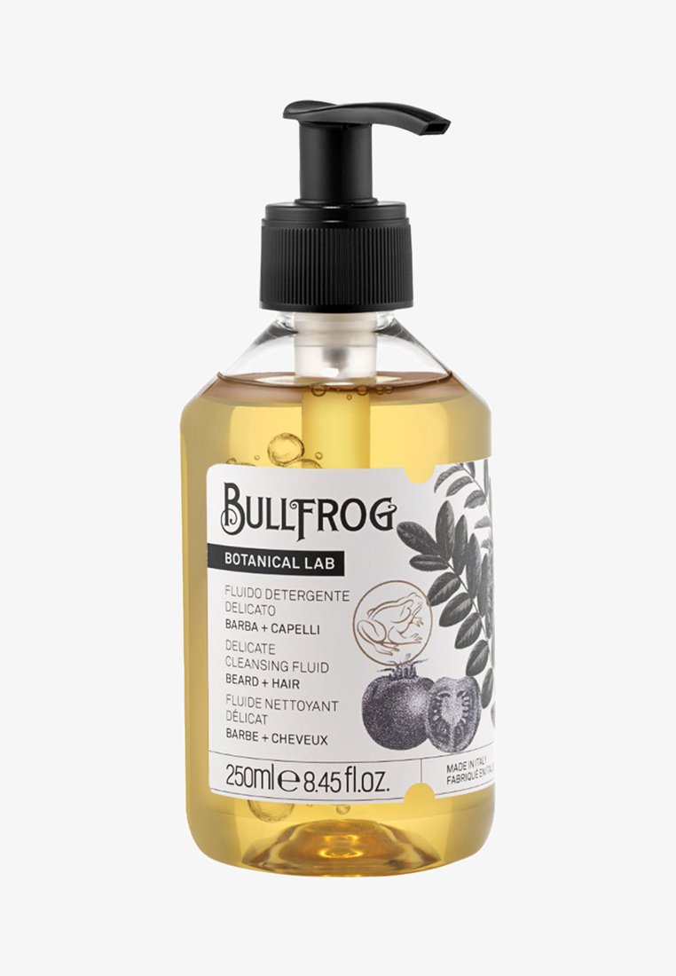 Bottiglia rotonda trasparente con un tappo a pompa nero. Contiene un fluido detergente giallo. Presenta un'etichetta bianca con testo nero e illustrazioni botaniche.