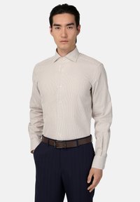 Camisa de hombre de manga larga con un patrón a rayas en color beige claro y burdeos. Tejido de algodón, cuello con botones, puños estándar, combinada con pantalones oscuros y un cinturón marrón.