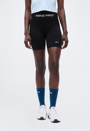 Persoon draagt zwarte Nike Pro short, blauwe Nike sokken, witte Nike sneakers en een lichtblauw shirt, staand tegen een effen witte achtergrond.