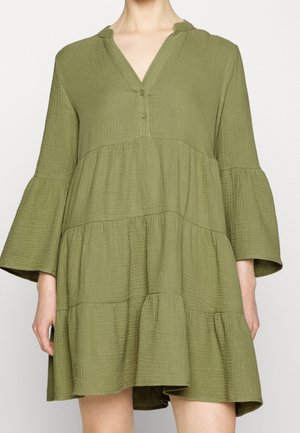 Kobieta w luźnej, warstwowej, oliwkowozielonej sukience z długimi, rozkloszowanymi rękawami i dekoltem w serek z dwoma guzikami.