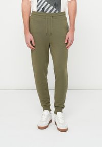 Muž v olivovo zelených teplákových nohaviciach a bielych teniskách stojaci pred bielym pozadím, ruky uvoľnene pozdĺž tela.