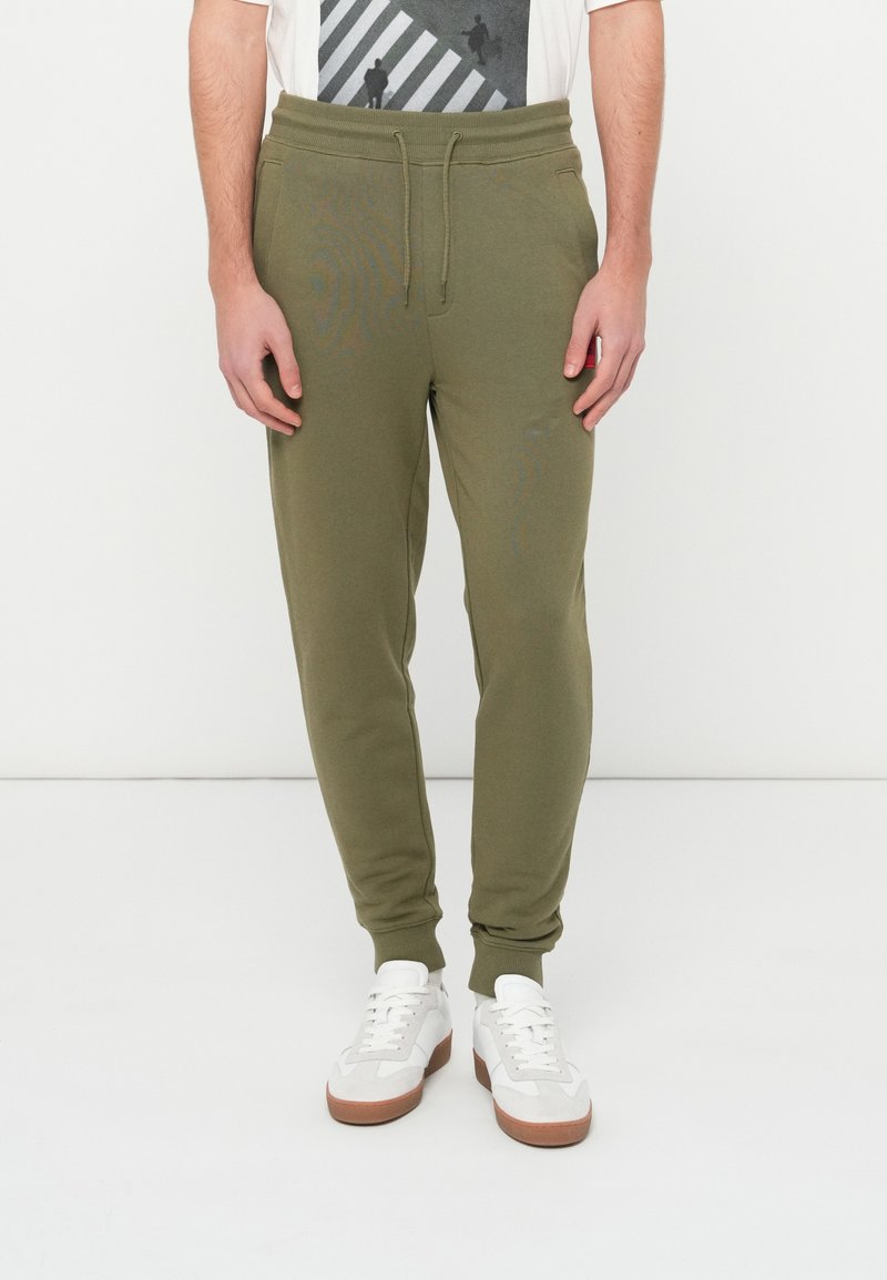 Muž v olivovo zelených teplákových nohaviciach a bielych teniskách stojaci pred bielym pozadím, ruky uvoľnene pozdĺž tela.