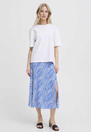 Femeie cu păr blond, purtând un top alb cu mâneci scurte, fustă albastră cu model și șliț, și sandale negre cu toc, pe un fundal simplu.