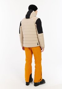 Beige gewatteerde jas met zwarte accenten, capuchon en elastische manchetten; gecombineerd met oranje waterdichte broek en zwarte skibotten.