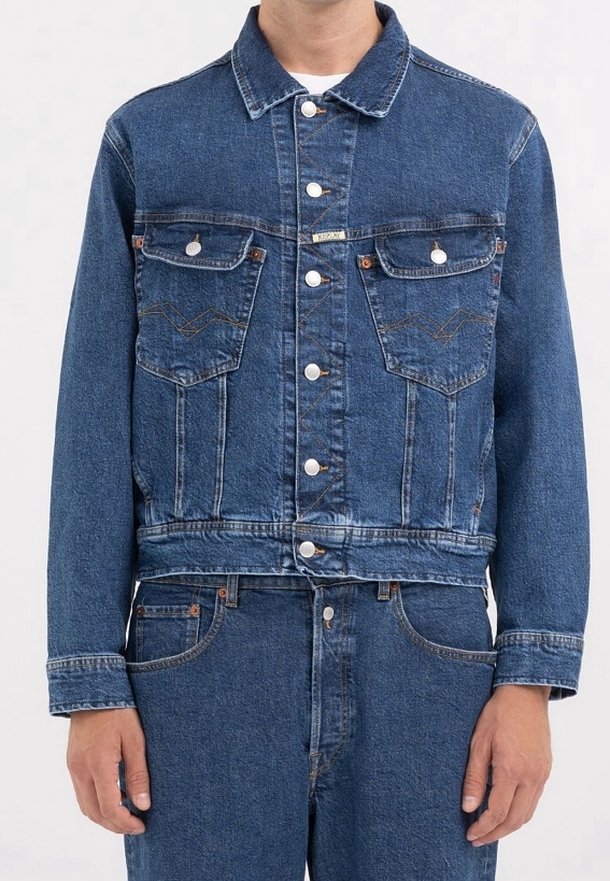 Blå denimjacka med knäppning framtill, två bröstfickor och en kortad design. Har synliga sömmar och metalldetaljer.