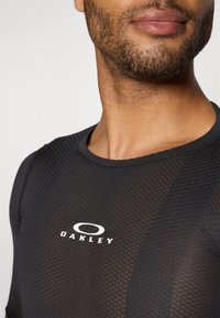 T-shirt de sport en maille noire avec une surface texturée, présentant un col rond et un logo "Oakley" blanc au niveau de la poitrine.