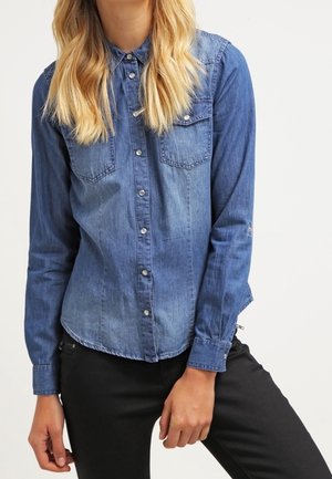Denim shirt in medium blue with long sleeves, button-down collar, two chest pockets, and a slightly curved hem. Black pants visible at the bottom.