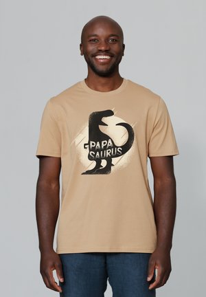 Beige katoenen t-shirt met een zwarte dino-afbeelding en de tekst "PAPA SAURUS". Korte mouwen met een ronde hals. Soepele stoftextuur.