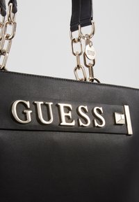 Sac à main noir Guess avec des anses en chaîne dorée et des lettres dorées du logo Guess à l'avant, avec une petite étiquette ronde Guess sur la sangle.